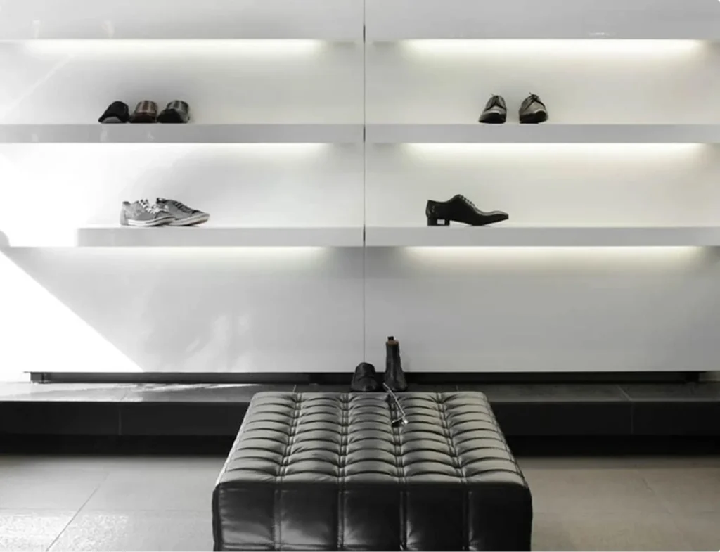 Présentation de différentes chaussures pour hommes sur des étagères minimalistes, avec un pouf noir au premier plan.
